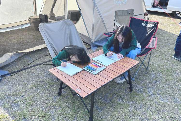 テント外のテーブルでスケッチブックに絵を描いている子ども2人